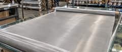 Stainless steel woven wire mesh roll opened on a factory worktable with packed rolls in the background
